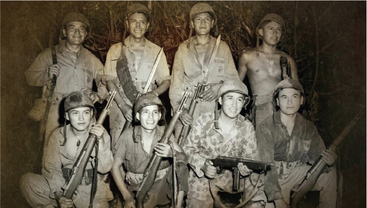 A sepia-toned historical photograph showing eight soldiers in military uniforms and gear during the Vietnam War. They are posed in two rows, holding rifles and other firearms, with some wearing helmets and others bareheaded. The background appears to be dense jungle foliage, suggesting a combat or field setting. The soldiers’ clothing includes standard-issue fatigues and camouflage patterns, and one individual is shirtless with a bandolier across the chest. The photo is from the documentary Native America: Vietnam Veterans