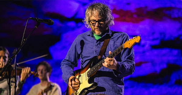 Musician playing an electric guitar on stage with a violinist in the background and blue-purple lighting.