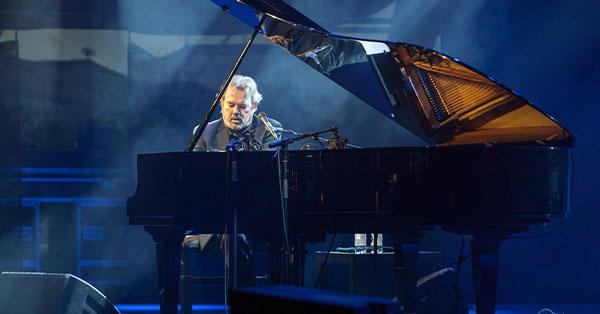 Musician playing a grand piano on stage with blue lighting and a microphone setup.