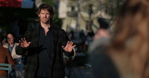 Man with hands raised in an outdoor setting, facing a blurred figure in the foreground.