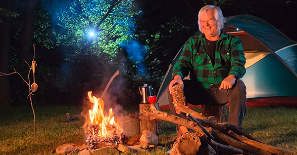 Smiling person by campfire with kettle and red cup; tent and trees visible in background.
