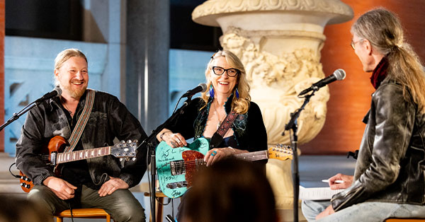 Two musicians with guitars sit and smile during an interview with a host in a decorative indoor setting.