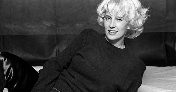 Black and white photo of a person with short, curly hair, wearing a dark sweater, reclining on a bed.