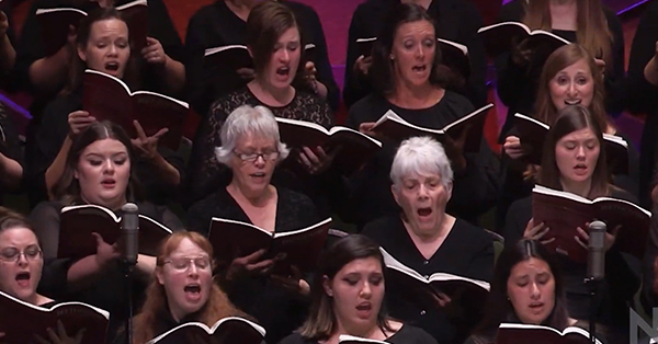 A choir of women singing from sheet music, wearing black attire, with microphones in front.
