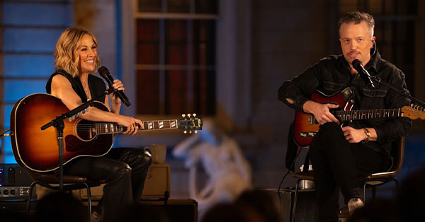 Two musicians seated, each holding a guitar and microphone, performing on stage with a blurred background.