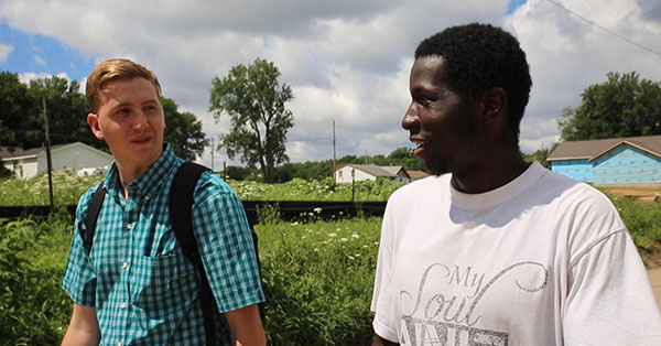 Two men walking outdoors, one in a plaid shirt with a backpack, the other in a white shirt. Greenery and houses in the background.