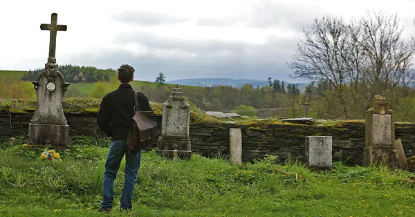 Man with a bag standing in a grassy cemetery, looking at old gravestones and a cross, with trees and cloudy sky in background.