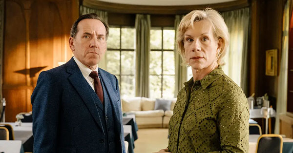 Two people standing in a well-lit room with large windows and green curtains, one in a suit and the other in a green shirt.