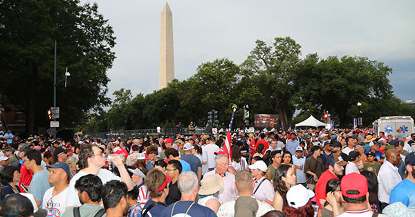 Crowd gathered near the Washington Monument, surrounded by trees and tents, with overcast sky.