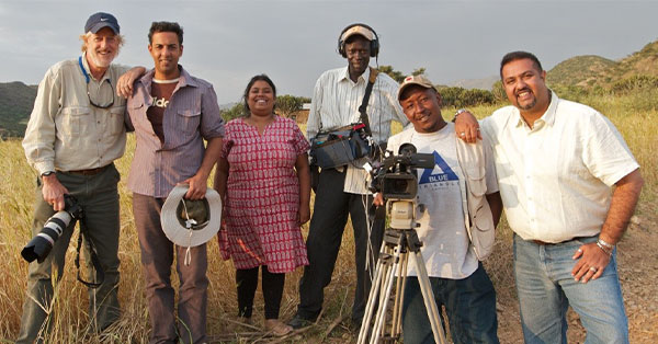 A group of six people with camera equipment stands in a field with hills in the background.