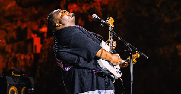 Musician passionately playing guitar on stage, with a microphone nearby and a colorful background.