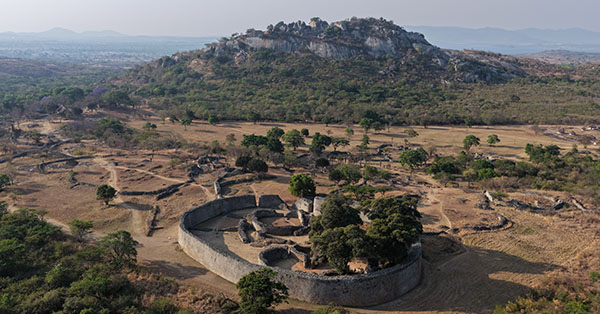Aerial view of ancient stone ruins and circular structures surrounded by trees with a rocky hill in the background.