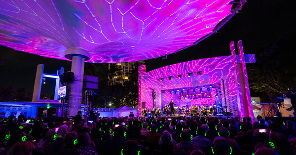 Outdoor concert with audience wearing headphones, stage lit in pink and purple lights, large canopy overhead.