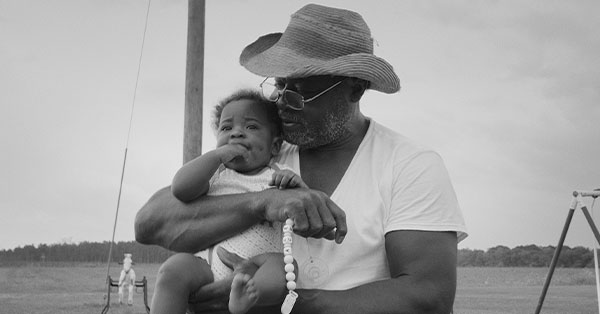 Man in a hat holding a baby outdoors with a field and overcast sky in the background.