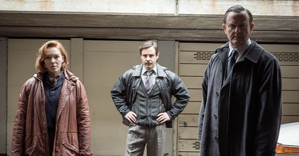 Three people standing in a garage, wearing coats and looking serious.