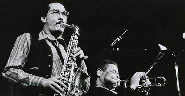 Two musicians performing; one plays a saxophone, the other a trumpet. Black and white photo.