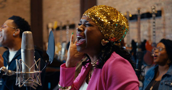 A woman in a pink jacket and gold hat sings passionately into a microphone with two people in the background.