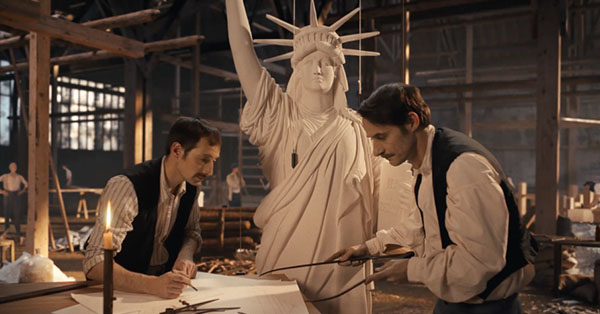 Two men working on a model of the Statue of Liberty inside a workshop with blueprints and a candle on the table.