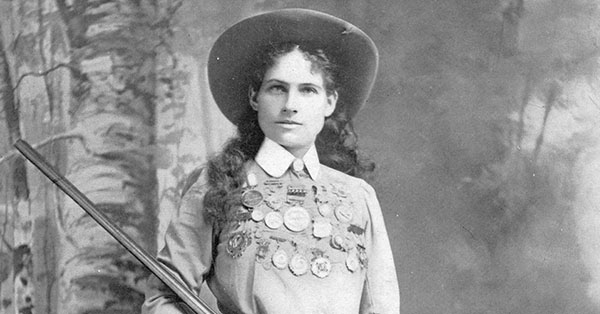 Woman in cowboy hat with medals on her shirt, holding a rifle, standing against a painted backdrop.