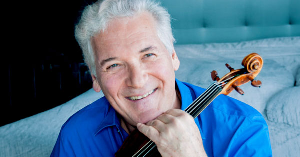 Man with gray hair in blue shirt smiling, holding a violin under his chin.
