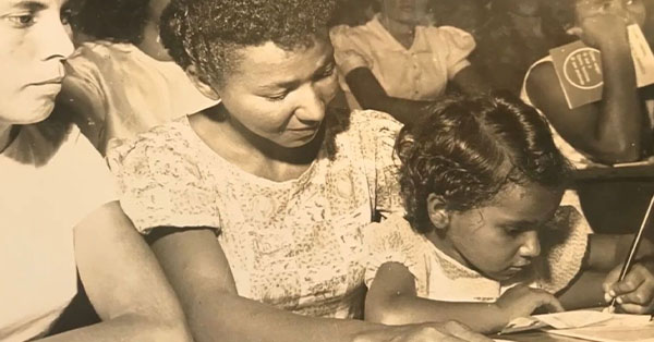 A child writes while sitting beside a woman in a crowded room. The scene is in sepia tone, suggesting a historical setting.