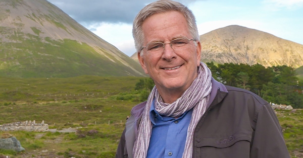 Rick Steves with a scarf standing in a green field with dirt covered mountains in the background.