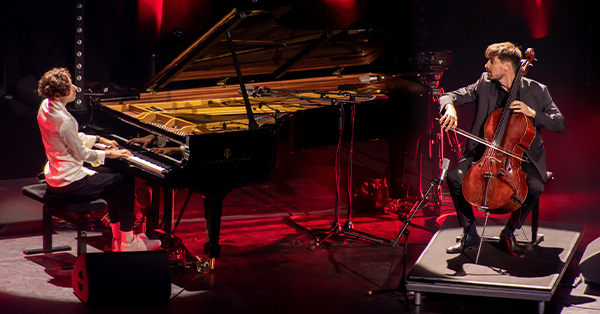 Pianist Peter Bence performs at a grand piano with cellist Luka Šulić during Peter Bence: Pianosphere in Paris. The two musicians face one another under warm red stage lighting.