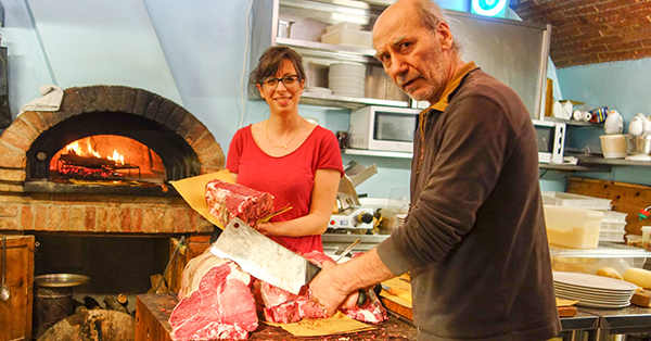 A butcher slices large cuts of beef on a wooden block while a woman stands beside him holding another cut of meat in a rustic kitchen. A wood-fired brick oven glows in the background.