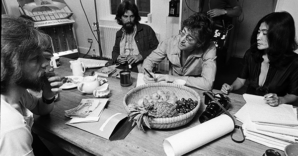  George Harrison, John Lennon and Yoko Ono working at a table with a couple of other individuals.