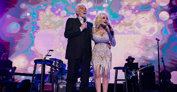 Kenny Rogers and Dolly Parton perform on Stage in front of a brightly colored background.