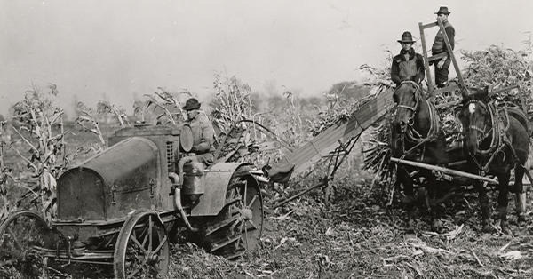 View from front of a man driving an International tractor pulling a corn picker. On the right two men stand on a horse-drawn wagon piled with cornstalks.