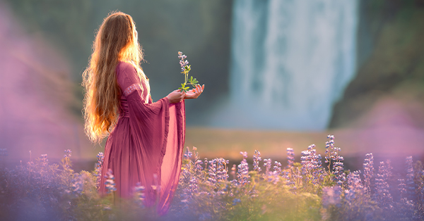 A blonde woman in a pink dress standing in a field of pink flowers with a waterfall in the background.