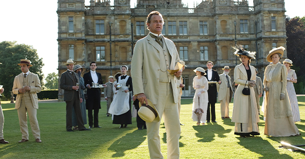 Hugh Bonneville as Lord Grantham standing on the lawn in front of Highclere Castle with many other characters from the show.