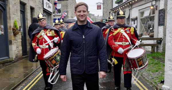 Nicholas Ralph walks down a English village street with a marching band behind him,