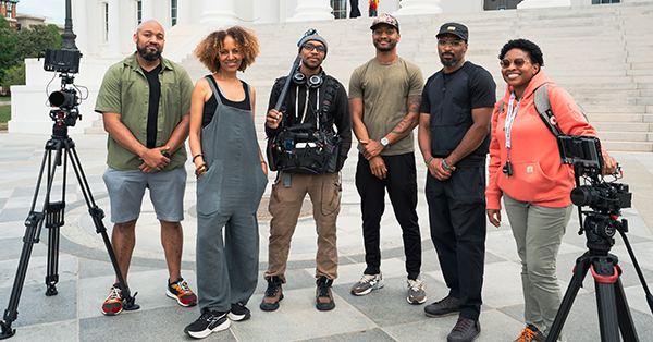 Six members of a film production crew stand together on the steps outside a large white government building. Two professional video cameras on tripods frame the group, and one crew member wears audio recording equipment.