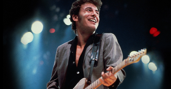Bruce Springsteen smiles while performing on stage, playing an electric guitar under bright concert lights. The blurred stage lighting in the background creates a lively live performance atmosphere.