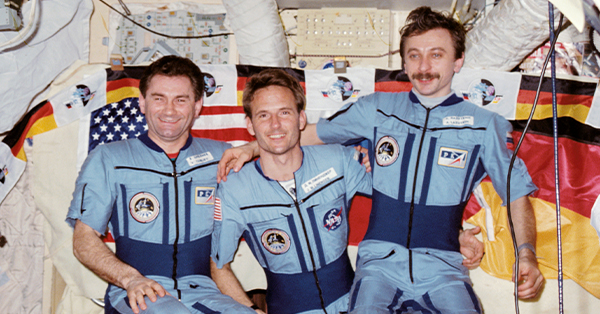 hree astronauts in matching light blue flight suits pose together inside a spacecraft, smiling with their arms around one another. U.S. and German flags hang behind them, highlighting an international space mission.