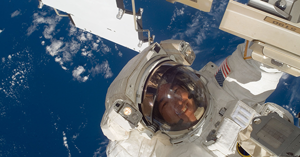 An astronaut in a white spacesuit smiles during a spacewalk outside the International Space Station, with Earth’s deep blue oceans and scattered white clouds stretching below. Parts of the station’s structure and solar panels frame the scene.