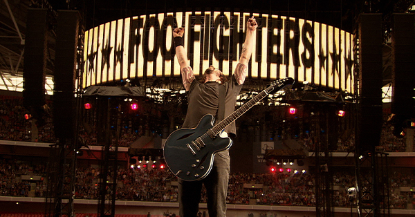 A guitarist fromthe Foo Fighters stands on a stadium stage with arms raised in celebration, an electric guitar strapped across his chest. A large illuminated screen displaying the band's name towers behind him as a packed crowd fills the venue.
