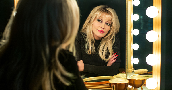 Promotional image of Dolly Parton looking into a lighted dressing room mirror. Her reflection is in sharp focus, with illuminated vanity bulbs and makeup containers visible in the foreground.