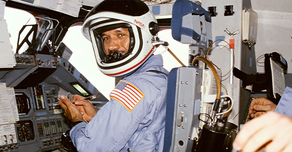 n astronaut wearing a NASA helmet and light blue flight suit sits at the controls inside a space shuttle cockpit, turning to smile at the camera. Flight instruments, switches, and control panels surround the astronaut, while another crew member's hand is partially visible in the foreground.