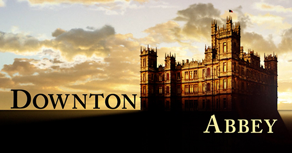 Promotional image for Downton Abbey featuring the grand estate at sunset beneath a dramatic sky. The series title appears across the foreground in large black and gold lettering.