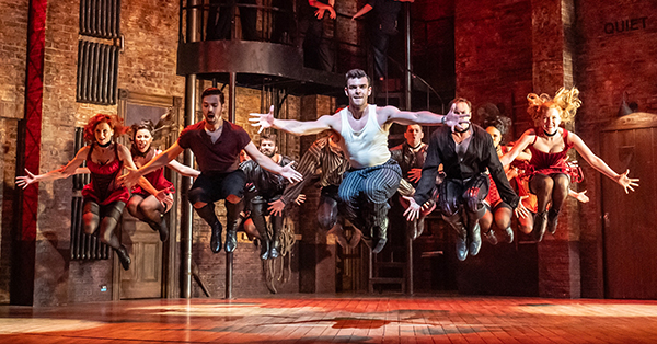 Kiss Me Kate dancers and performers leap into the air in perfect unison during a high-energy stage production. The industrial-style set, warm theatrical lighting, and dynamic choreography create a dramatic performance scene.