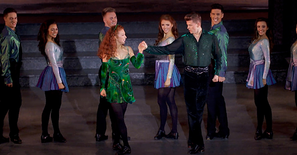 Dancers perform a traditional Irish dance during Riverdance 25th Anniversary Show. A lead couple dances at center stage while other performers in colorful costumes smile and pose behind them.