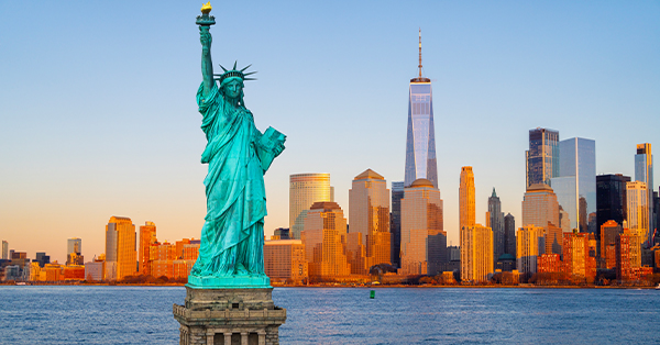 The Statue of Liberty stands in the foreground with the Lower Manhattan skyline behind it at sunset. One World Trade Center rises above the surrounding buildings, with warm golden light reflecting across the harbor.