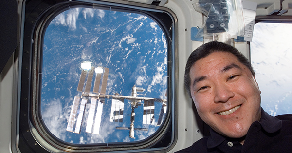 A smiling astronaut poses inside the International Space Station beside a window overlooking Earth. Through the window, another section of the space station with its solar arrays is visible against the blue oceans and white clouds below.