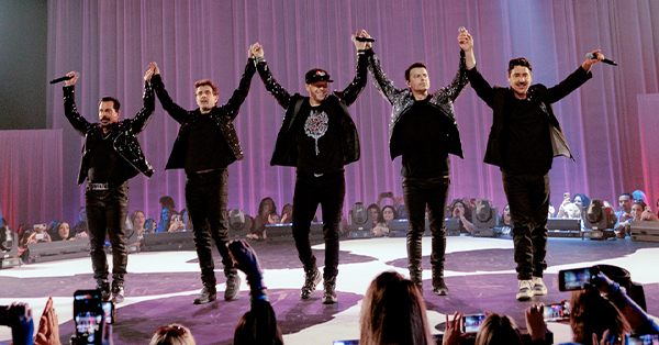 The five members of New Kids on the Block stand on stage with joined hands raised in celebration during New Kids on the Block: iHeartRadio Live. Fans surround the stage as purple lighting fills the arena.