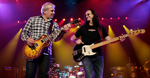 Guitarist Alex Lifeson and lead singer, bassist, and keyboardist Geddy Lee perform together on stage during a live concert. Colored stage lights illuminate the musicians as they smile and play their instruments.